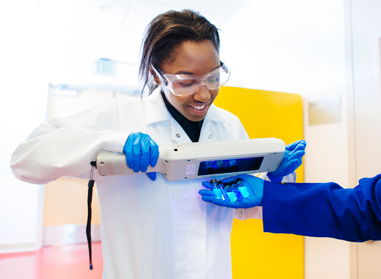 Student in lab coat shining a black light on florescent test tubes.