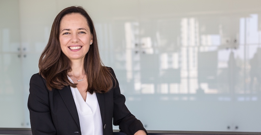 Professor Noemi Petra in a white shirt and black jacket poses in front of a reflective, opaque glass wall.