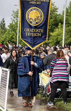 Trevor Albertson ('09), now the dean of Lassen Community College, bears the graduate division flag for the School of Social Sciences, Humanities and Arts at the spring 2019 commencement.